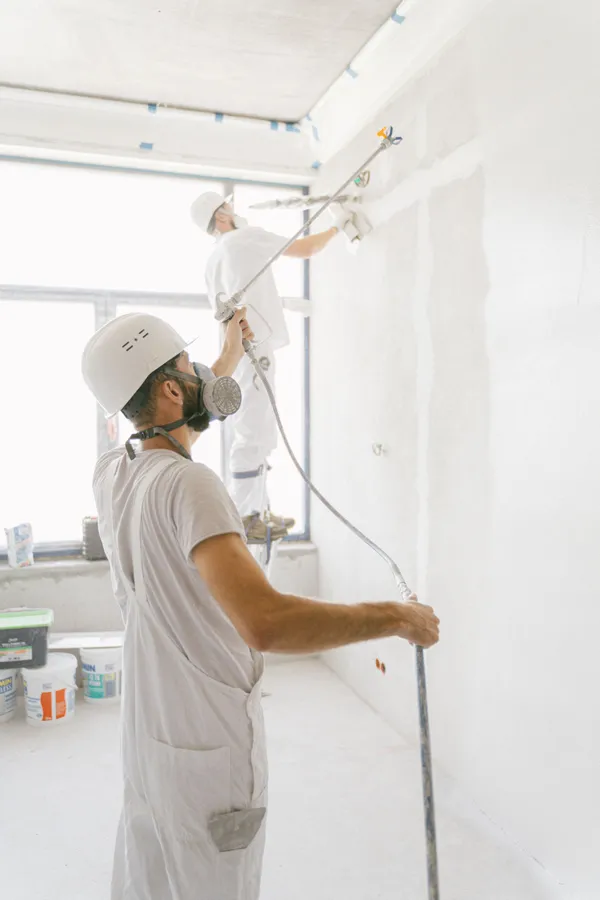Two painters in protective equipment preparing and coating interior walls beside large windows